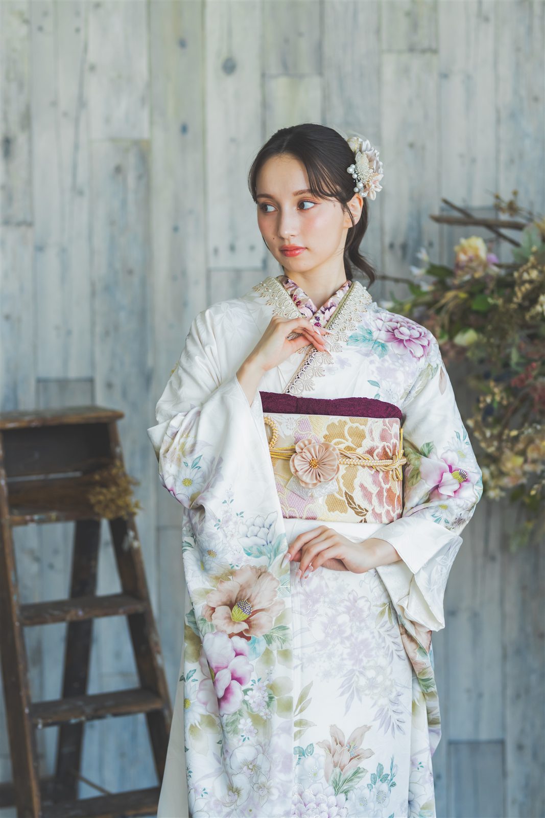 白地に淡い花の振袖を着て、花と木の背景の前に立つ上半身