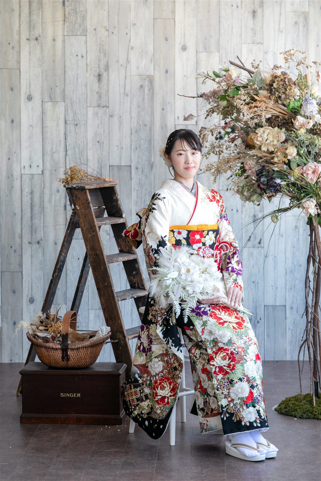 花と木のセットの前で、黒地に花の振袖を着て立つ様子
